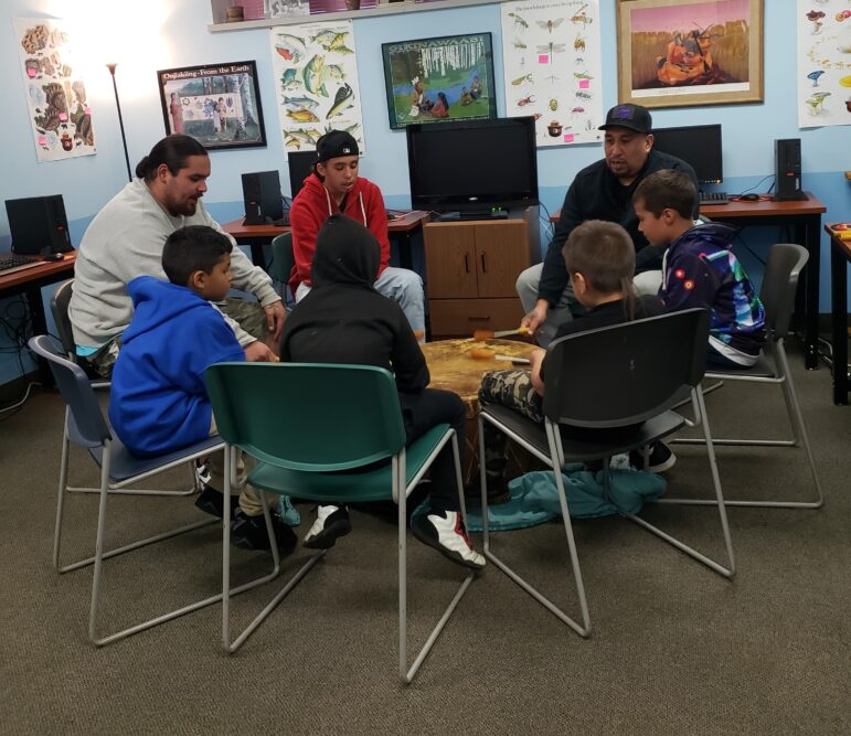 A Native American man teaches a group of young boys gathered around a large drum in a classroom.