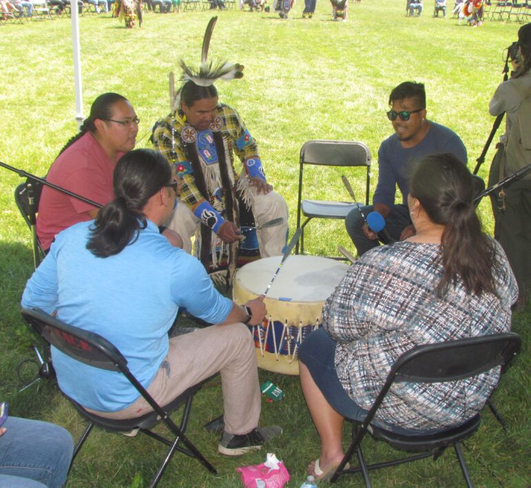 Native American men and women sit around a powwow drum outdoors. One man is in traditional regalia.
