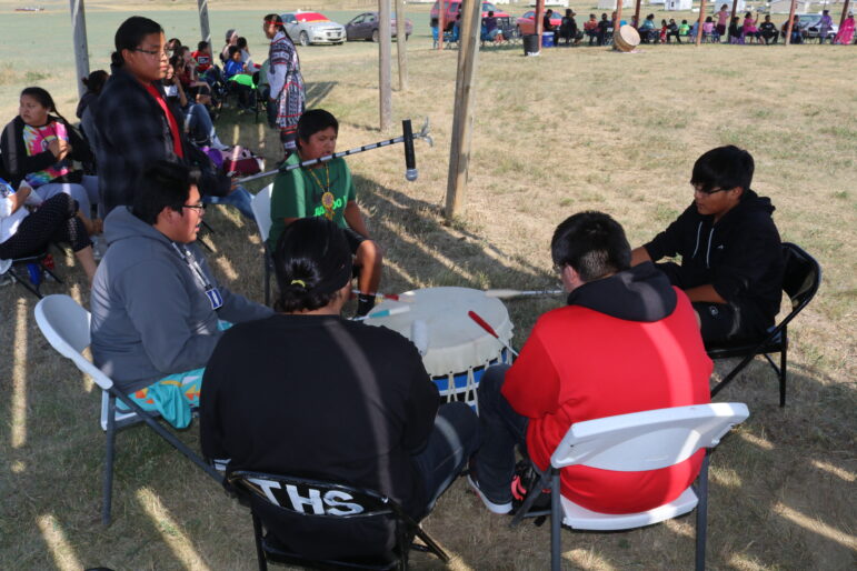 A group of Native American men and boys sitting in a circle around a large powwow drum outdoors.