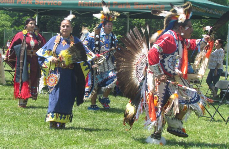 Native American men and women dancing in traditional regalia at a powwow, with feather fans and colorful beadwork on display.