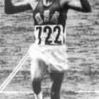 Billy Mills (USA 722) crosses the finish line with arms raised, winning the 10,000m gold medal at the 1964 Tokyo Olympics.