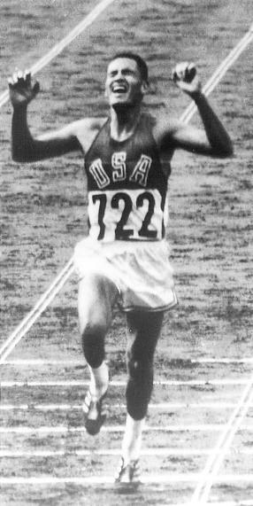 Billy Mills (USA 722) crosses the finish line with arms raised, winning the 10,000m gold medal at the 1964 Tokyo Olympics.