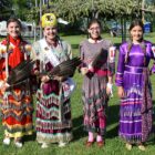 Four young Native American women posing in traditional regalia, called Jingle Dresses, at an outdoor event.