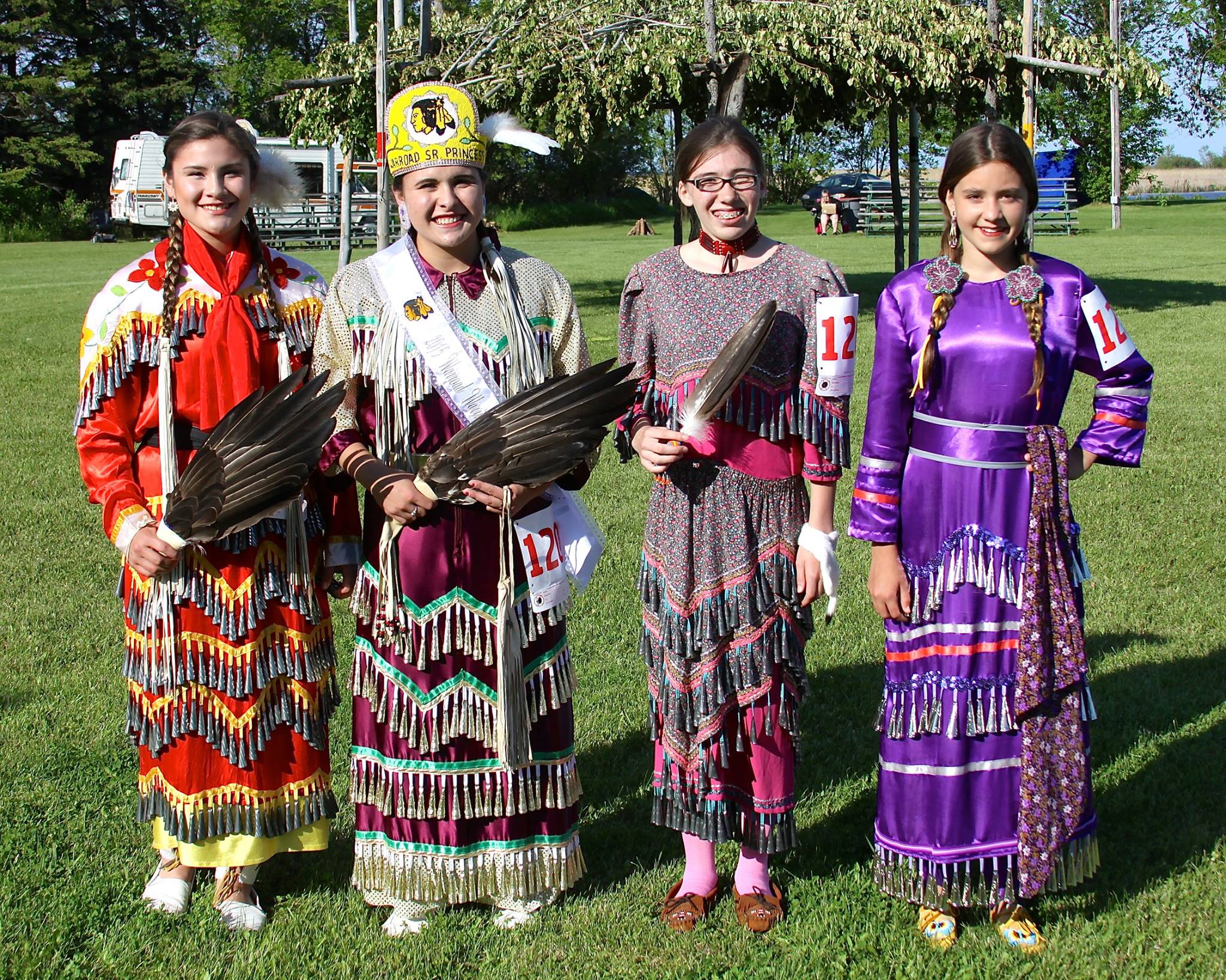 Four young Native American women posing in traditional regalia, called Jingle Dresses, at an outdoor event.