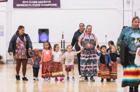 Native youth and elders in traditional ribbon skirts at a cultural gathering.