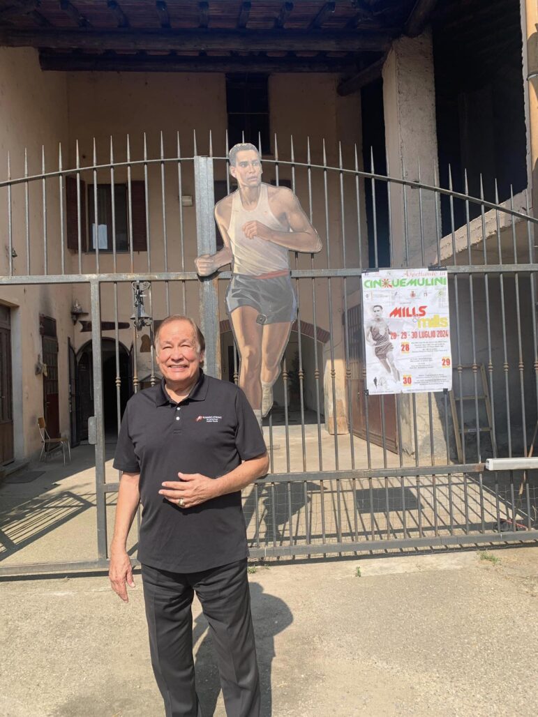 Billy Mills visits Italy for the Cinque Mulini event, posing in front of a gate with a cutout of his younger self running.