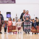 Native American children in colorful ribbon skirts hold hands with elders at a cultural event in a school gym.