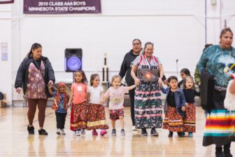 Native American children in colorful ribbon skirts hold hands with elders at a cultural event in a school gym.