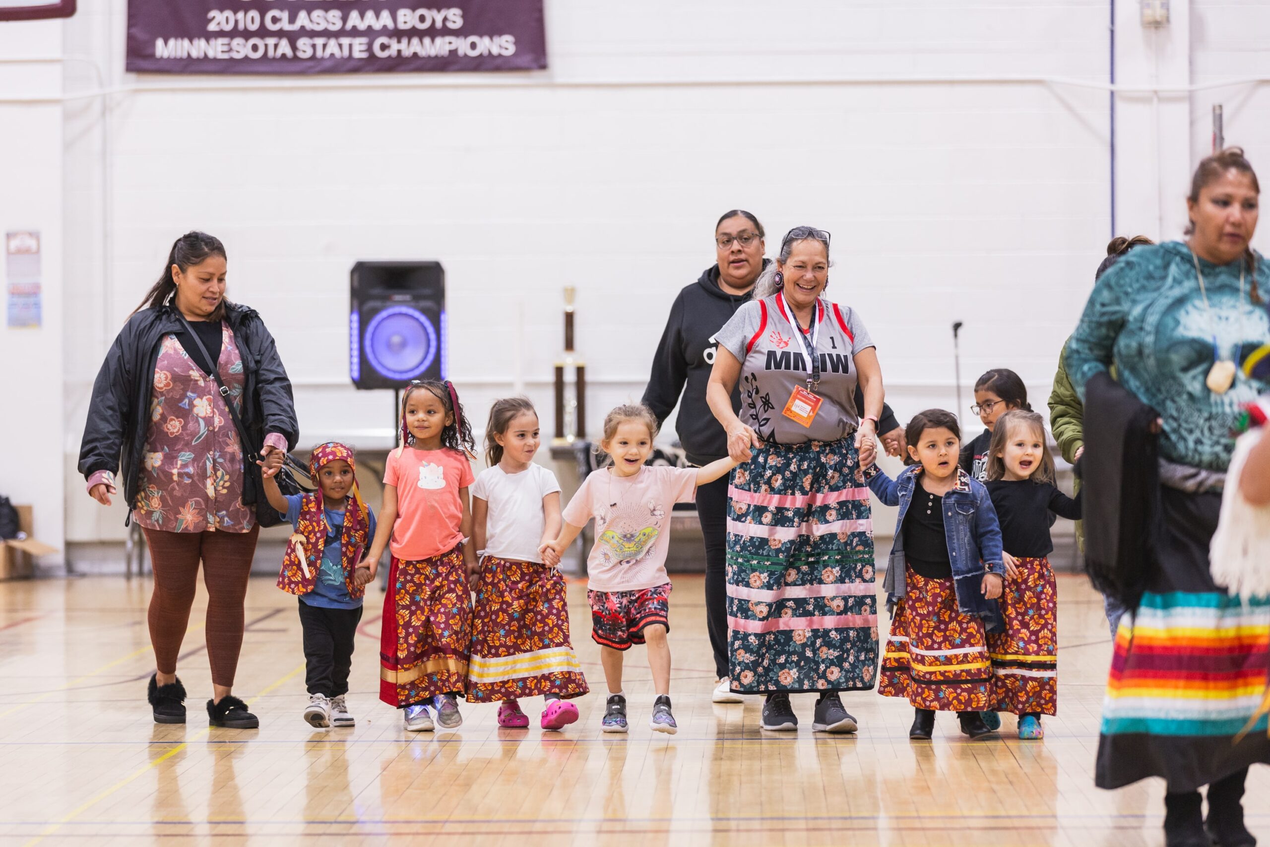 Native American children in colorful ribbon skirts hold hands with elders at a cultural event in a school gym.
