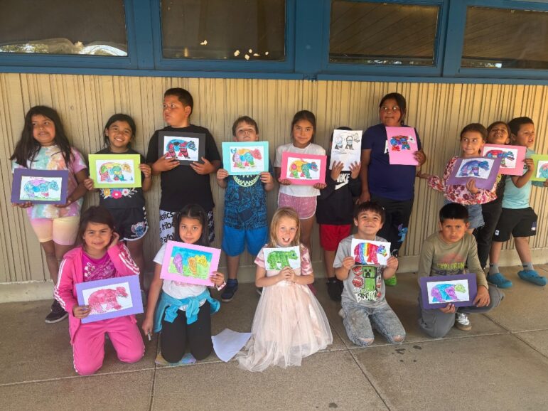 A class of smiling students holds up colorful bear paintings from an art project by Dreamstarter Teacher Serena Inda.