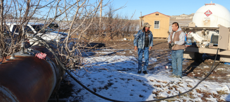 Two men stand in a snowy yard during a propane delivery, providing heat for a family via the Winter Warmth program.