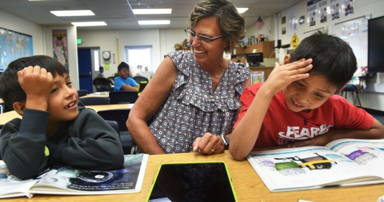 A smiling teacher helps two young Native students with reading in a classroom, showing the Running Strong education program.