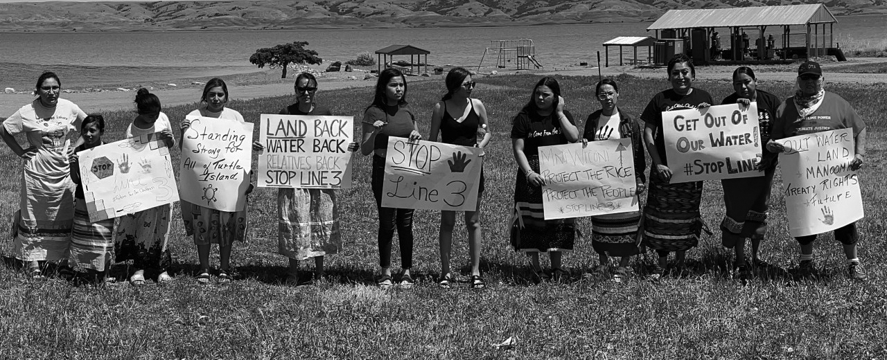 Indigenous women water protectors protest, holding signs like "STOP Line 3" to support Native youth-led environmental action.