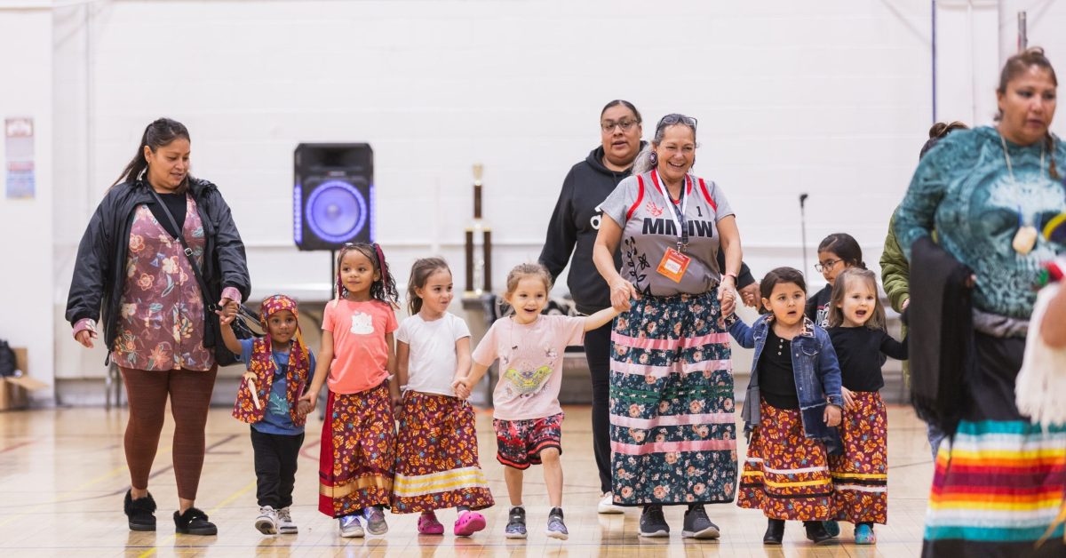 Native American children in colorful ribbon skirts hold hands with elders at a cultural event in a school gym.