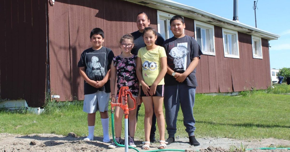 Native american children and adult stand beside new water spigot outside rural home